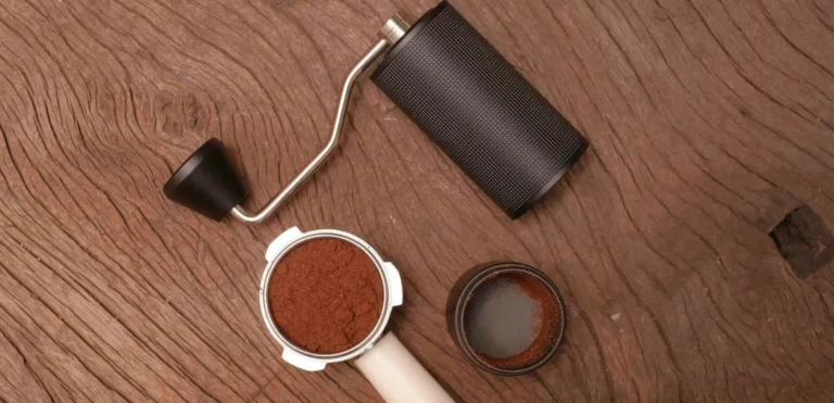 Visão superior em uma mesa mostrando um moedor de café manual cilíndrico preto. Ao lado, há um porta-filtro de máquina de café espresso cheio de café moído e o compartimento inferior do moedor com restos de pó de café.